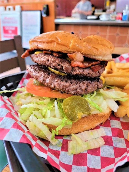 A towering burger with two patties, bacon, lettuce, tomato, pickles, and a bun, served with fries on the side.