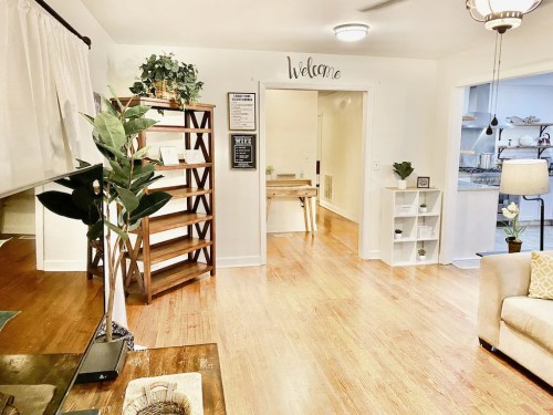 Cozy interior with wooden floors, a welcome sign, plants, and open spaces leading to a bright kitchen area.