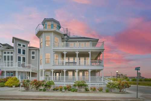 A large, multi-level beach house with balconies, surrounded by colorful flowers, against a pink sunset sky.