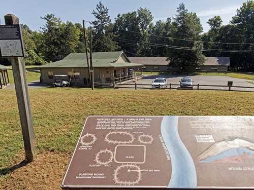 A sign and informational display in front of a building, surrounded by trees and parked cars.