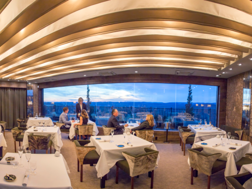 Elegant restaurant interior with diners enjoying a view of the sunset through large windows. Soft lighting and modern decor.