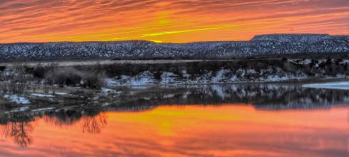 A vibrant sunset reflects on a calm river, with snow-covered hills in the background and colorful clouds above.