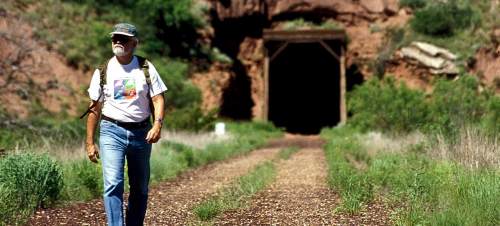 A man walks along a trail leading to a dark tunnel, surrounded by greenery and rocky terrain.