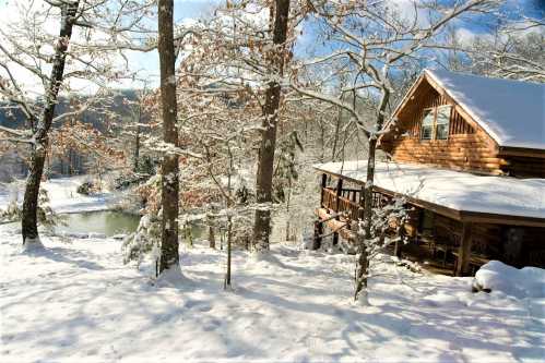 A cozy log cabin surrounded by snow-covered trees and a serene lake, under a bright blue sky.