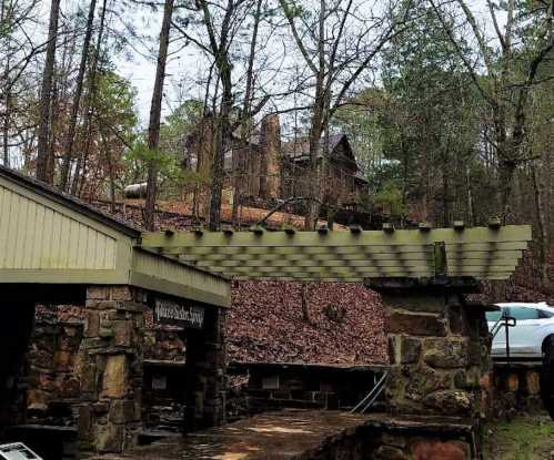 A stone structure with a wooden pergola, surrounded by trees and a hillside house in a wooded area.