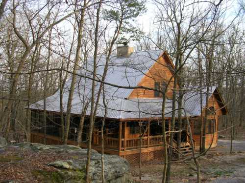 A rustic wooden cabin surrounded by bare trees and rocky terrain, featuring a sloped roof and a porch.