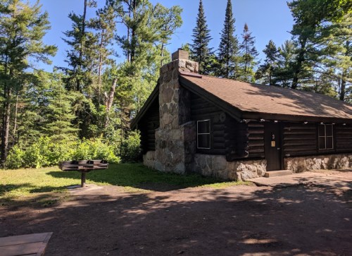 A rustic log cabin surrounded by trees, featuring a stone chimney and a nearby grill area.