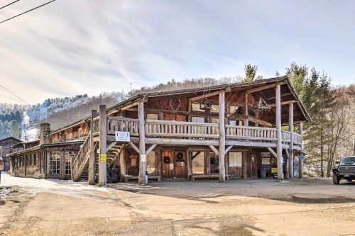 A rustic wooden building with a balcony, surrounded by trees and mountains, under a clear sky.