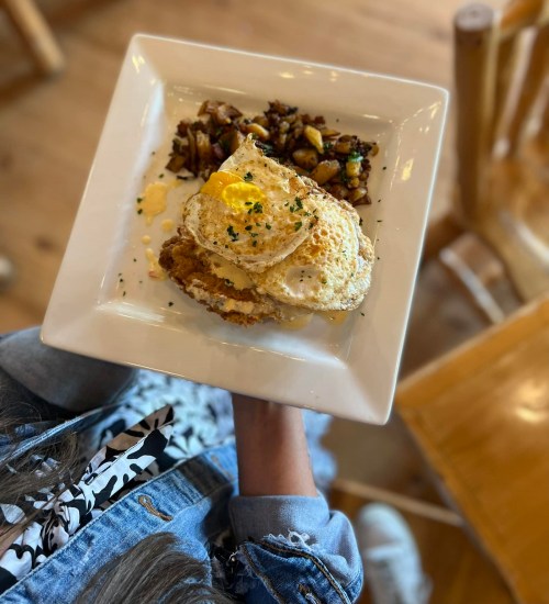 A person holding a plate with two fried eggs, seasoned potatoes, and a pat of butter, set on a wooden table.