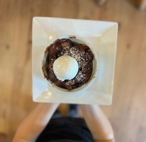 A person holds a square dish with warm chocolate dessert topped with a scoop of vanilla ice cream and powdered sugar.