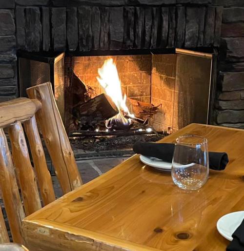 A cozy restaurant setting with a wooden table, black napkin, glass of water, and a warm fireplace in the background.