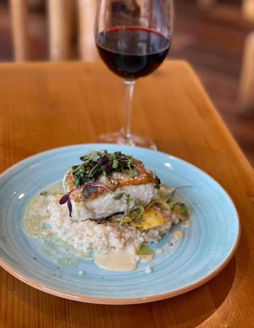 A plate of fish topped with microgreens, served on a bed of creamy rice, alongside a glass of red wine.