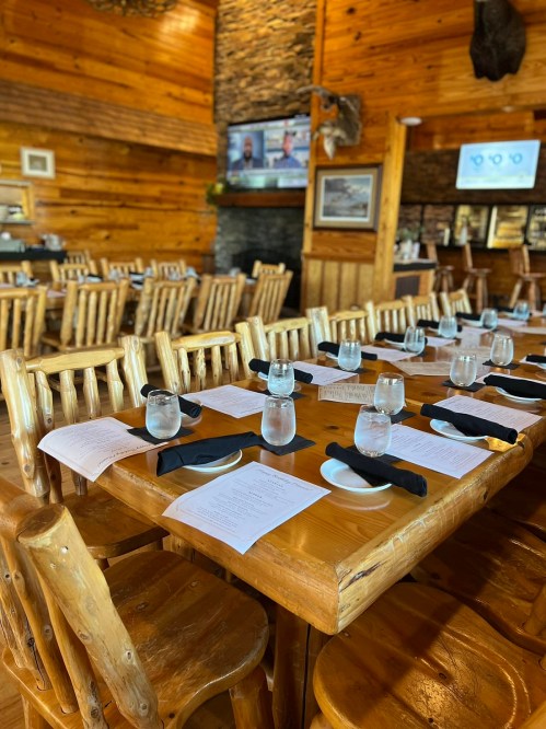 A rustic restaurant interior with wooden tables set for dining, featuring menus, water glasses, and a stone fireplace.