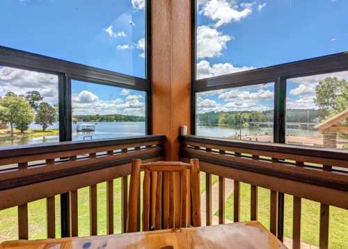 View from a cabin window overlooking a lake, with blue skies and fluffy clouds, and a wooden chair in the foreground.