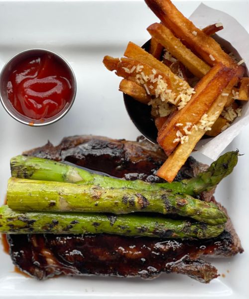 A plate featuring grilled steak topped with asparagus, served with fries and a small cup of ketchup.