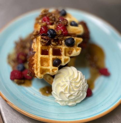 A golden waffle topped with berries, nuts, and syrup, served with a dollop of whipped cream on a blue plate.