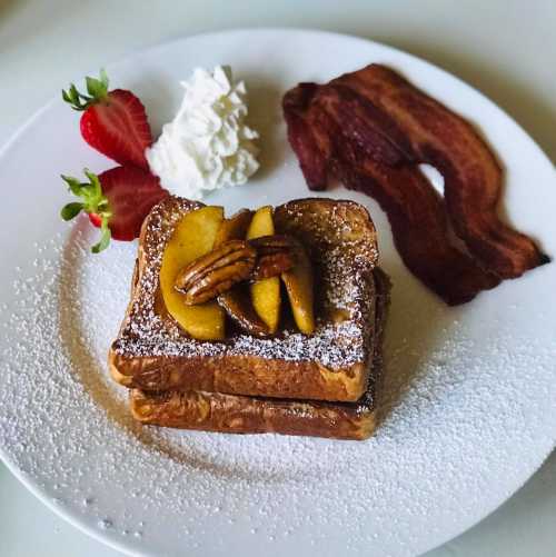 A plate of French toast topped with apples and pecans, served with strawberries, whipped cream, and crispy bacon.