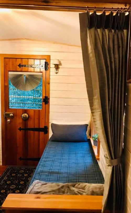 Cozy interior of a small room featuring a single bed, wooden door, and curtains, with a decorative window.