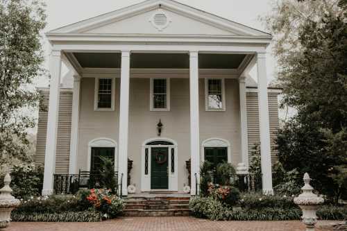 A large, elegant house with white columns, green door, and lush gardens in front.