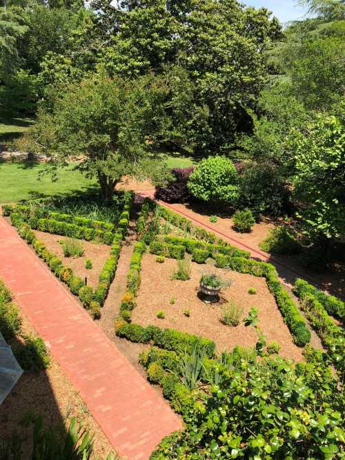 Aerial view of a landscaped garden with geometric flower beds, pathways, and lush greenery surrounding a central fountain.