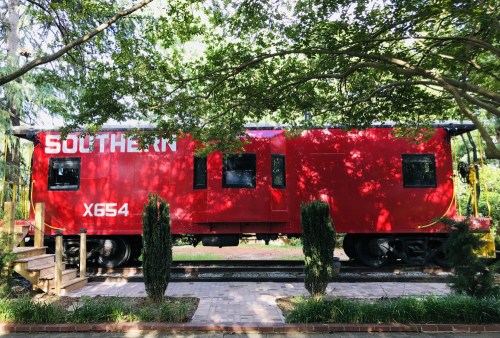 A red Southern Railway caboose parked under trees, with steps leading up to it on a brick path.