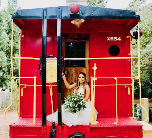 A bride in a white dress sits on a red train car, holding a bouquet of greenery, smiling at the camera.