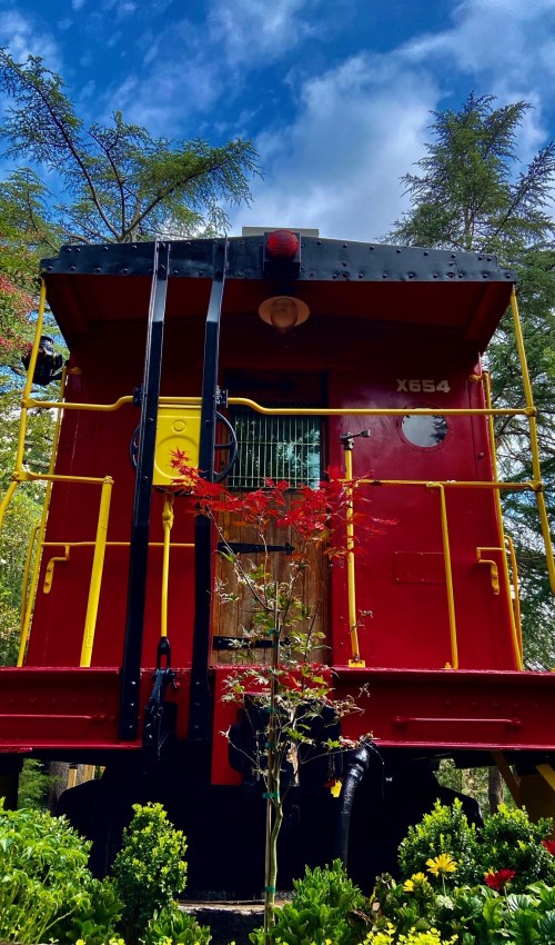 A red train caboose stands among greenery under a blue sky, with a small tree in front.