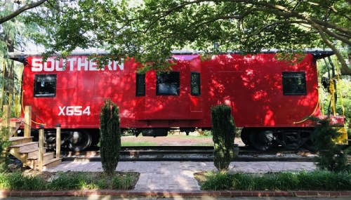 A red Southern Railway caboose parked under trees, with steps leading up to it on a brick path.