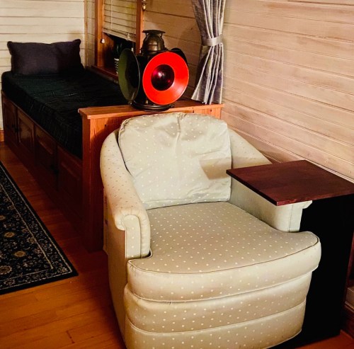Cozy room featuring a bed, a green armchair, and a vintage gramophone on a side table. Warm wooden walls.