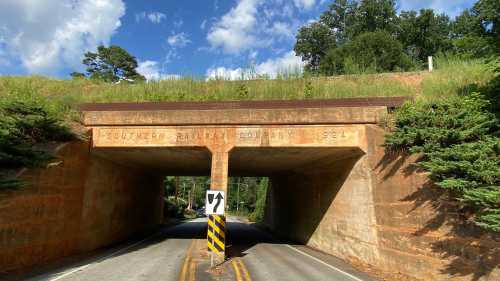 A weathered railway overpass with "Southern Railway Company 1924" engraved, above a two-lane road surrounded by greenery.