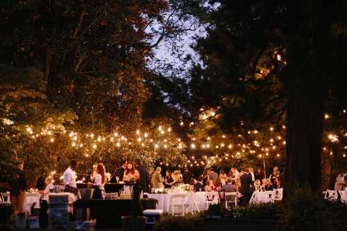 A beautifully lit outdoor event with guests dining at tables under string lights amidst trees.