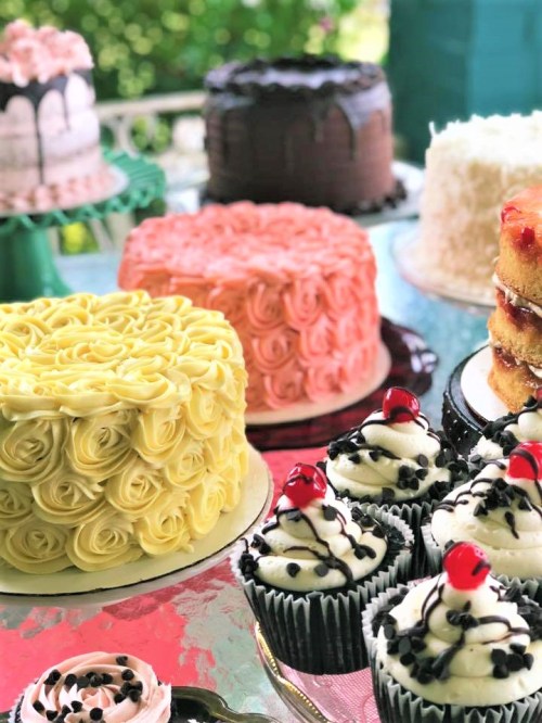 A variety of beautifully decorated cakes and cupcakes in pastel colors, featuring floral designs and cherries on top.