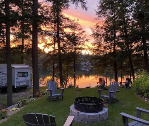 A serene lakeside scene at sunset, featuring a fire pit surrounded by chairs and trees reflecting on the water.