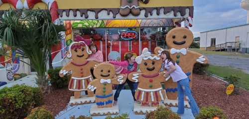 Four people pose playfully with large gingerbread figures outside a colorful candy-themed shop.