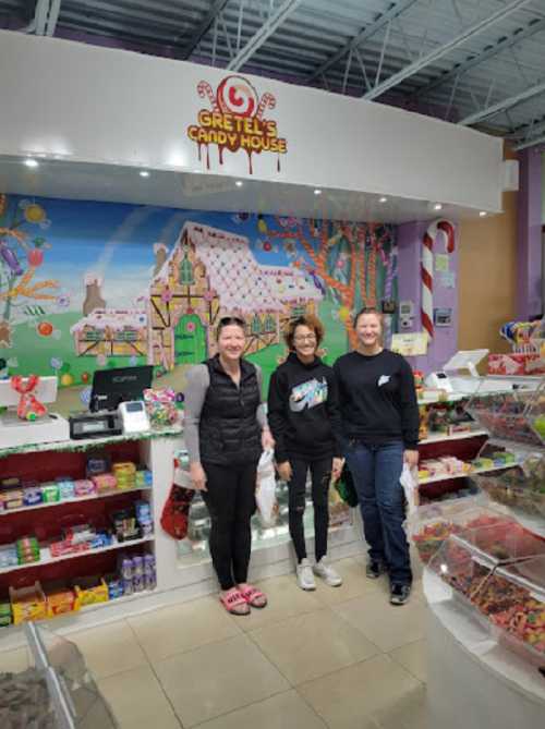 Three people stand in front of a colorful candy display at Gretel's Candy House, smiling and holding bags of candy.