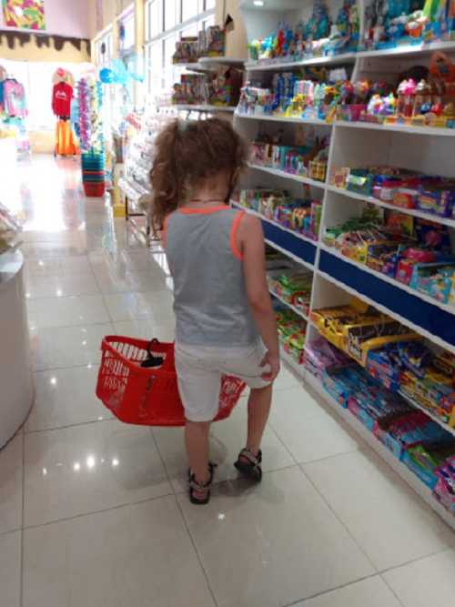 A child with curly hair walks through a colorful store, holding a shopping basket. Shelves are filled with toys and treats.