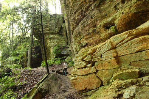 A person walking along a trail beside colorful rock formations and lush greenery in a forested area.
