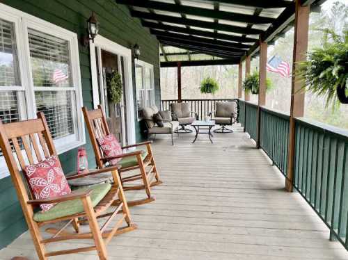 A cozy porch with rocking chairs, cushions, and a table, surrounded by greenery and small American flags.