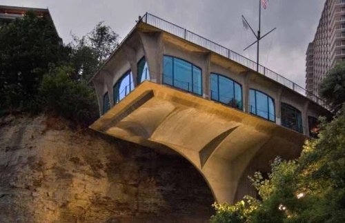 A modern, cantilevered building with large windows, perched on a cliff, surrounded by greenery and a cloudy sky.