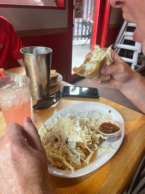 A person holds a taco filled with shredded cabbage, with a plate of food and a drink on the table.
