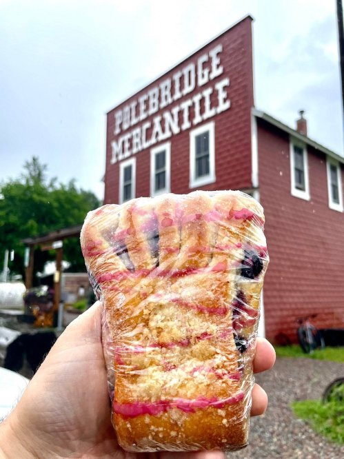 A hand holding a wrapped pastry in front of the red Polebridge Mercantile building.