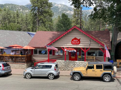 A cozy restaurant with a red roof, outdoor seating, and American flags, surrounded by trees and mountains.