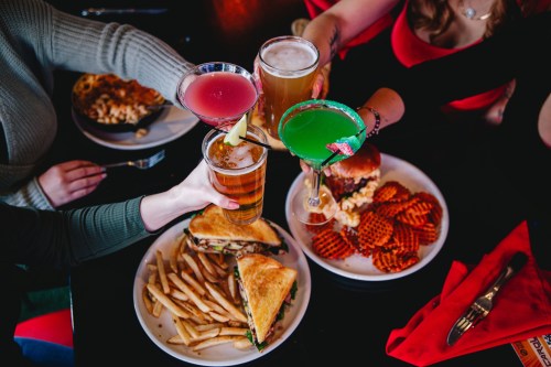 A group toasting with colorful drinks over a table filled with sandwiches, fries, and appetizers.