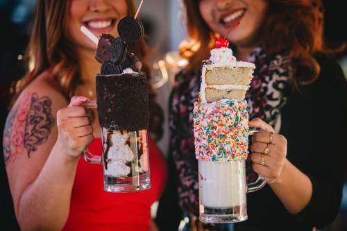 Two women hold oversized milkshakes topped with cake, sprinkles, and cookies, smiling in a vibrant setting.
