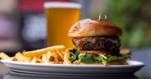 A juicy burger with cheese, lettuce, and tomato, served with fries and a glass of beer in the background.