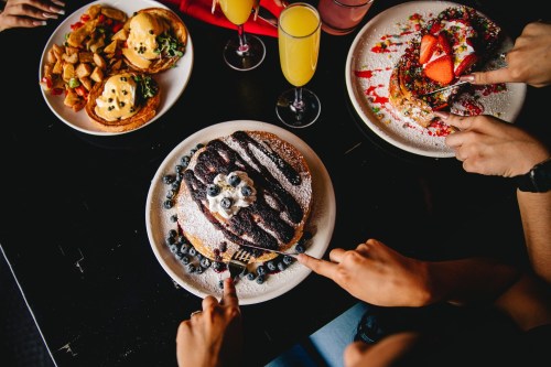 A table with colorful brunch dishes, including pancakes topped with blueberries and chocolate, and mimosas.
