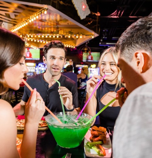 A group of friends enjoying a colorful drink from a large bowl, sharing straws in a lively bar setting.