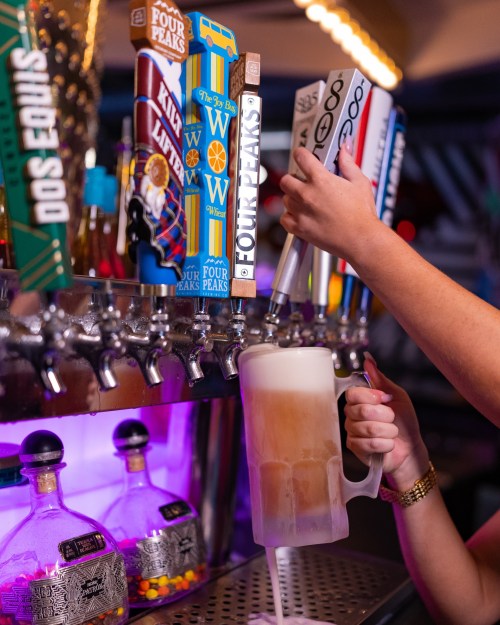 A person pours beer from colorful taps into a frosty mug, with bottles of liquor visible in the background.