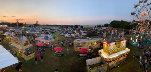 A vibrant fair scene at sunset, featuring food stalls, a Ferris wheel, and crowds enjoying the festivities.