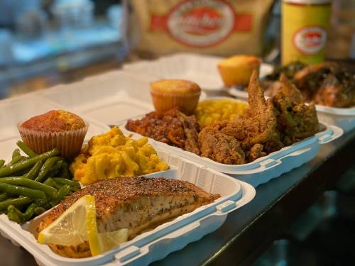 A takeout meal featuring seasoned fish, fried chicken, macaroni, green beans, cornbread muffins, and baked beans.